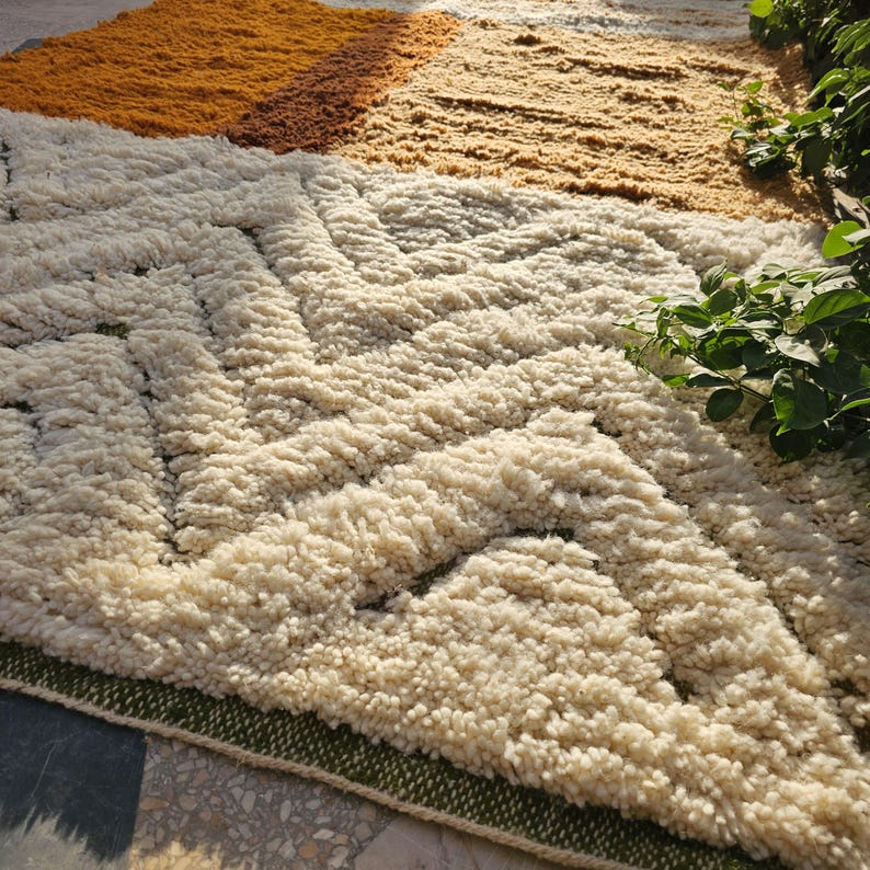 May include: A textured, geometric rug with a cream, tan, and orange color scheme. The rug features a repeating pattern of cream-colored shapes against a tan background, with a border of green. The rug is made of a soft, plush material.