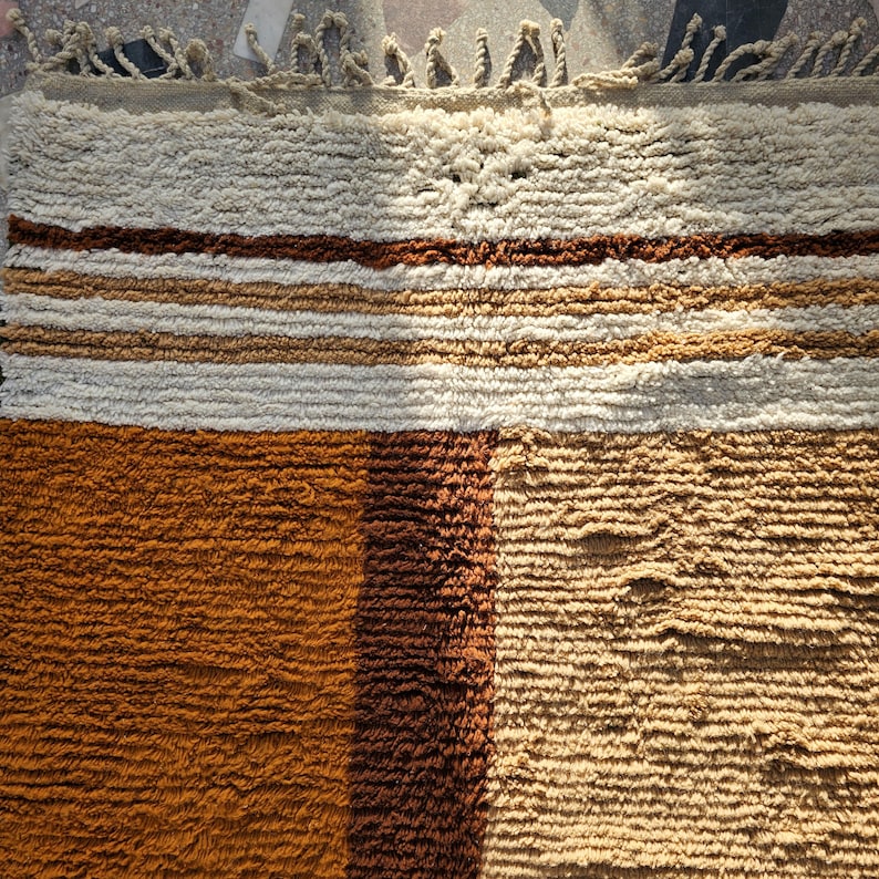 May include: A textured, rectangular rug with a mix of colors. The rug features sections of cream, brown, and tan, with horizontal stripes and a fringed edge. The rug's design is a blend of geometric and organic elements.