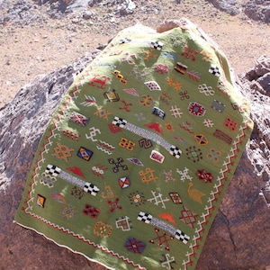 May include: A green, rectangular textile with colorful geometric patterns and a white border. The textile is draped over a large rock, with a mountainous background. The patterns include squares, zigzags, and other shapes in red, black, and yellow.