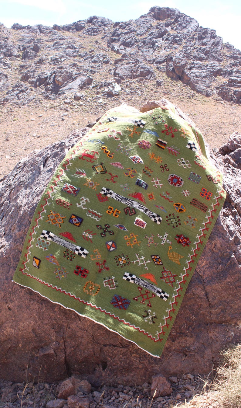May include: A green, rectangular textile with colorful geometric patterns and a white border. The textile is draped over a large rock, with a mountainous background. The patterns include squares, zigzags, and other shapes in red, black, and yellow.