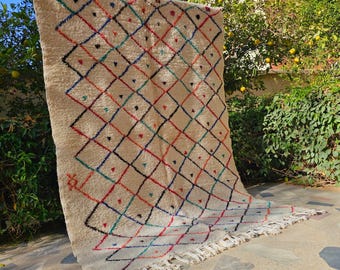 May include: A beige rug with a diamond pattern in red, blue, green, and black. The rug has a fringe along the bottom edge and is displayed outdoors. The diamond pattern is made up of small triangles and dots.
