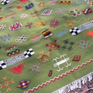 May include: A green, hand-woven rug with colorful geometric patterns. The rug features a variety of embroidered shapes in red, orange, yellow, and blue. Two black and white checkered bands are placed on the rug. The rug has a white fringe.