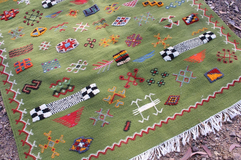 May include: A green, hand-woven rug with colorful geometric patterns. The rug features a variety of embroidered shapes in red, orange, yellow, and blue. Two black and white checkered bands are placed on the rug. The rug has a white fringe.