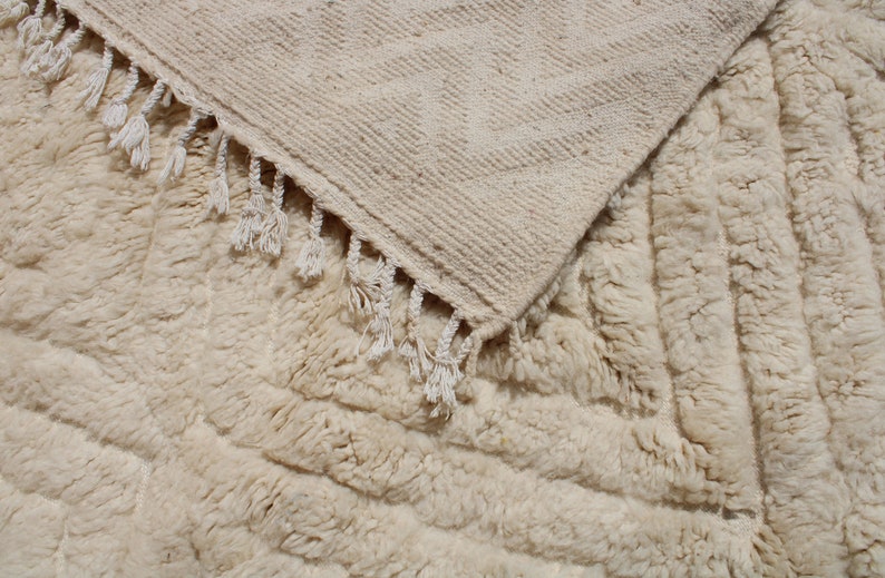 Puede incluir: Dos alfombras tejidas de color crema con flecos. La alfombra superior tiene un patr&oacute;n geom&eacute;trico y la alfombra inferior tiene una superficie texturizada y esponjosa.
