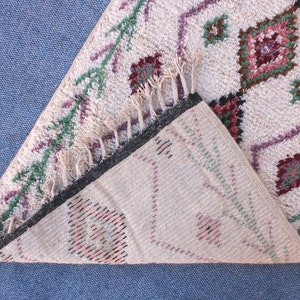 May include: A white woven rug with a geometric pattern in shades of pink, green, and brown. The rug has fringe on all sides and is folded in half.