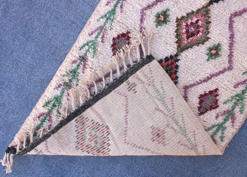 May include: A white woven rug with a geometric pattern in shades of pink, green, and brown. The rug has fringe on all sides and is folded in half.