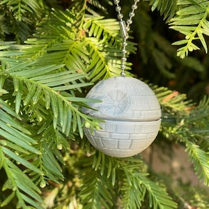Könnte beinhalten: Ein weißer, runder Weihnachtsbaumschmuck in Form des Todessterns aus Star Wars, der an einer silbernen Kette an einem grünen Weihnachtsbaum hängt.