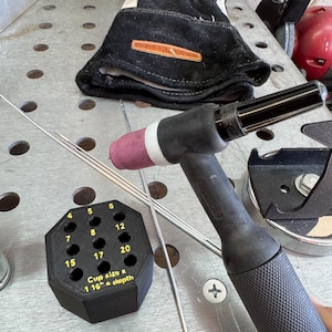 May include: Welding tools and equipment, including a black and gray TIG torch, welding gloves, and a black octagonal holder with numbered holes. The holder has text that reads "Cup Kizu & 1 16" a depth". Several silver welding rods are also present.