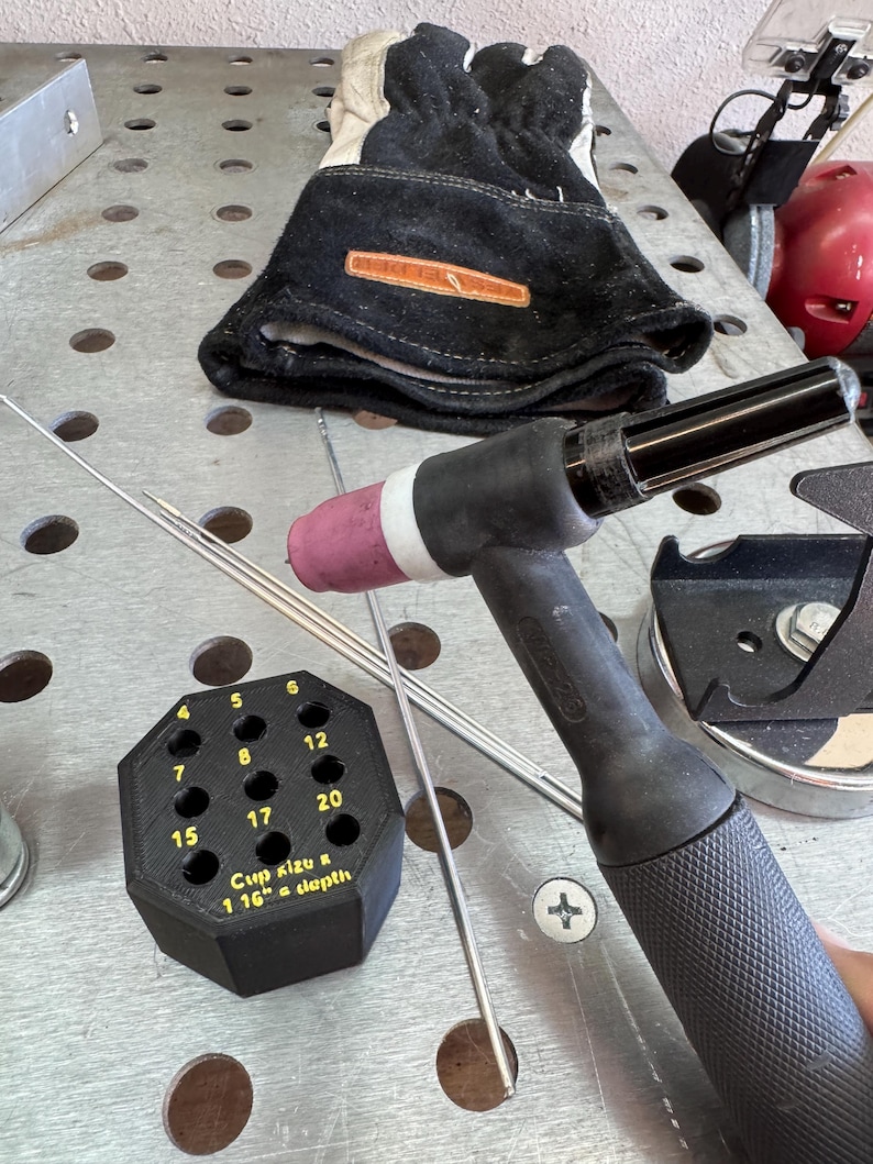 May include: Welding tools and equipment, including a black and gray TIG torch, welding gloves, and a black octagonal holder with numbered holes. The holder has text that reads "Cup Kizu & 1 16" a depth". Several silver welding rods are also present.
