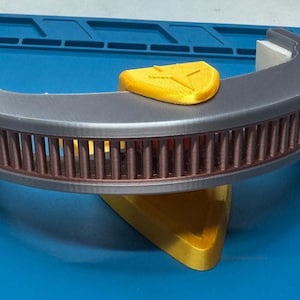 May include: A 3D-printed object with a curved, gray top section featuring vertical brown bars. A yellow accent piece sits on top, and the object rests on a yellow triangular base. The background is a blue surface.