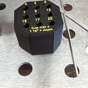 May include: A black, multi-holed welding cup size holder with the numbers 4, 5, 6, 7, 8, 12, 15, 17, and 20 printed in yellow. The text "Cup Kizu R 1 16" a depth" is also visible. Several metal rods are next to the holder.
