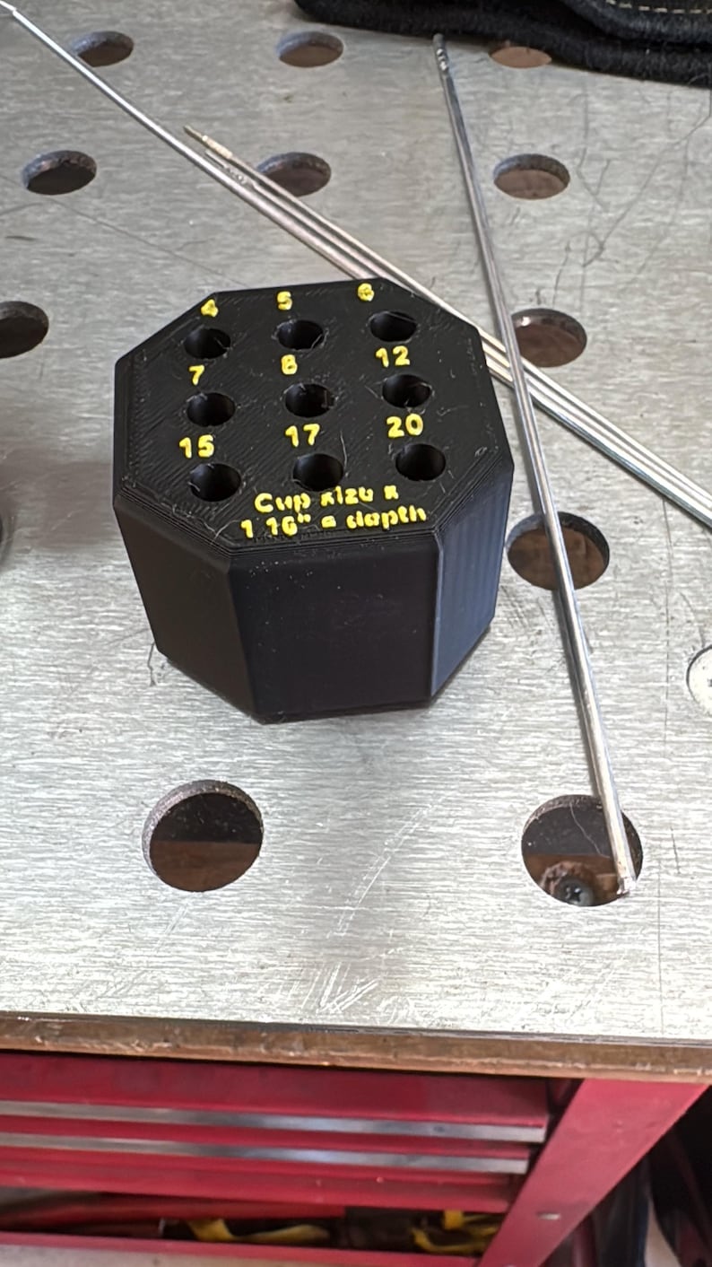 May include: A black, multi-holed welding cup size holder with the numbers 4, 5, 6, 7, 8, 12, 15, 17, and 20 printed in yellow. The text "Cup Kizu R 1 16" a depth" is also visible. Several metal rods are next to the holder.