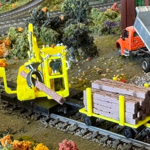 May include: A yellow model train excavator holding a wooden beam, alongside a flatbed train car carrying wooden beams. An orange dump truck is visible in the background.