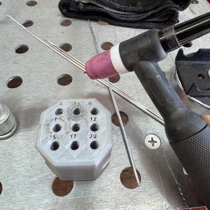May include: A close-up of welding tools and accessories. A black and silver TIG torch with a pink ceramic nozzle is positioned near a gray, multi-holed holder. Several silver welding rods are visible, along with a black welding glove.