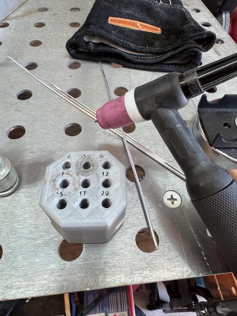 May include: A close-up of welding tools and accessories. A black and silver TIG torch with a pink ceramic nozzle is positioned near a gray, multi-holed holder. Several silver welding rods are visible, along with a black welding glove.