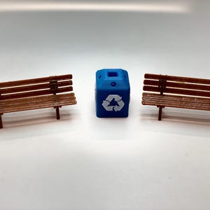 May include: A miniature park scene featuring two brown wooden benches and a blue recycling bin with a white recycling symbol. The benches have a slatted design and are supported by brown legs.