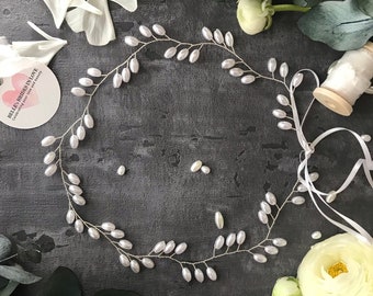May include: A delicate silver headband with small white pearl-like beads arranged in a vine-like pattern. The headband is on a gray surface with white flowers and a spool of white ribbon.
