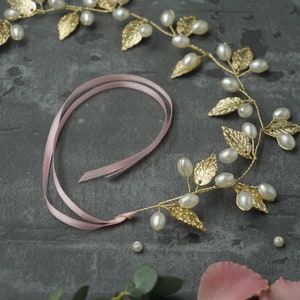 May include: A gold leaf and pearl hair vine with a pink ribbon. The vine is made of gold wire and has small gold leaves and white pearls. The ribbon is tied in a bow at the end of the vine.