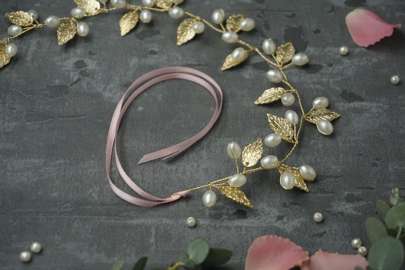May include: A gold leaf and pearl hair vine with a pink ribbon. The vine is made of gold wire and has small gold leaves and white pearls. The ribbon is tied in a bow at the end of the vine.