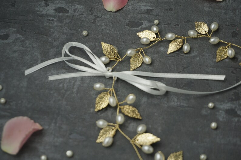 May include: A gold leaf and pearl hair vine with a white satin ribbon. The vine is made of delicate gold leaves and pearls, and the ribbon is tied in a bow.