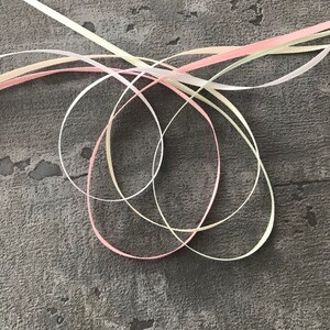 May include: Three satin ribbons in pink, cream, and light green are intertwined and knotted together on a gray textured surface.