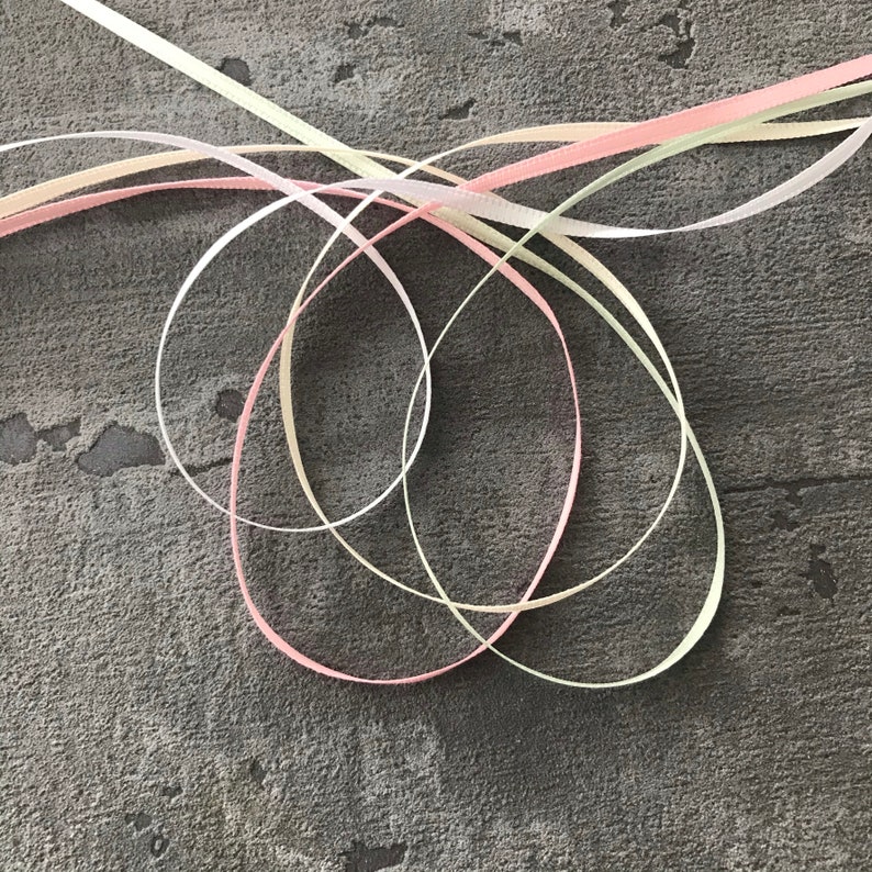 May include: Three satin ribbons in pink, cream, and light green are intertwined and knotted together on a gray textured surface.