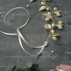 May include: A gold leaf and pearl hair vine with a white satin ribbon. The vine has small gold leaves and white pearls. The ribbon is tied in a knot.