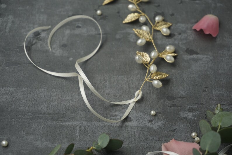 May include: A gold leaf and pearl hair vine with a white satin ribbon. The vine has small gold leaves and white pearls. The ribbon is tied in a knot.