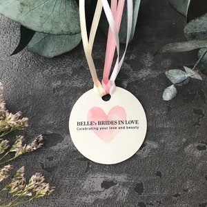 May include: A round, white tag with a pink watercolor heart and the text "Belle's Brides in Love" and "Celebrating your love and beauty".