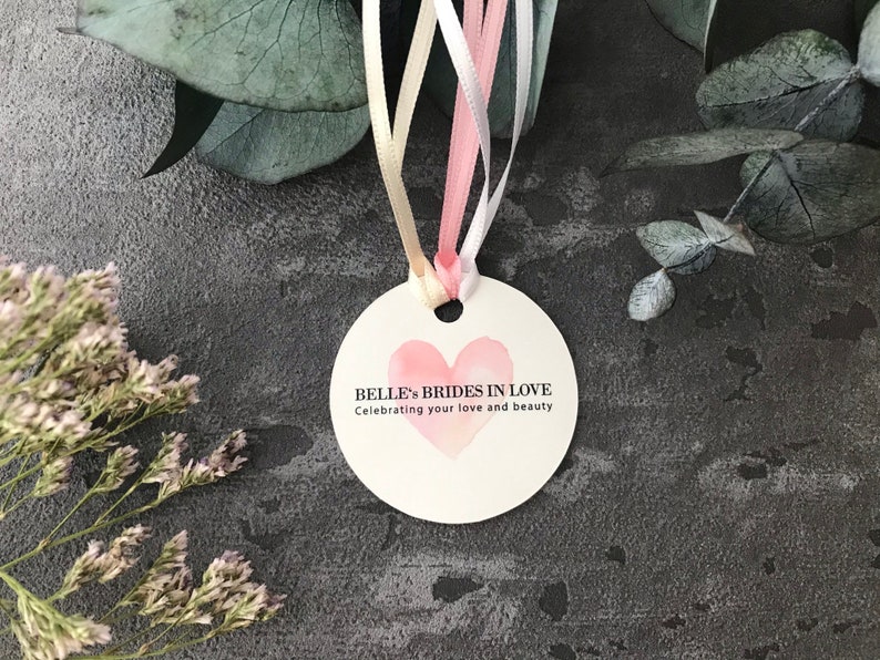 May include: A round, white tag with a pink watercolor heart and the text "Belle's Brides in Love" and "Celebrating your love and beauty".