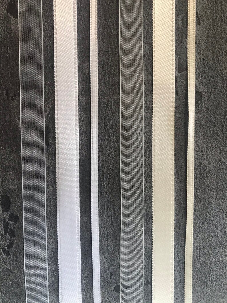 May include: Assortment of white and ivory ribbon trims, including sheer and satin, displayed against a dark gray background.