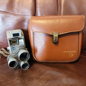 May include: Vintage Keystone 8mm movie camera and a matching brown leather case. The camera is silver and black with the name "Keystone" printed on it. The case has a metal clasp and the Keystone logo.