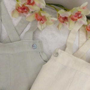 May include: Two pairs of linen overalls, one in a light green color and the other in a cream color. Both pairs have adjustable straps and button closures.