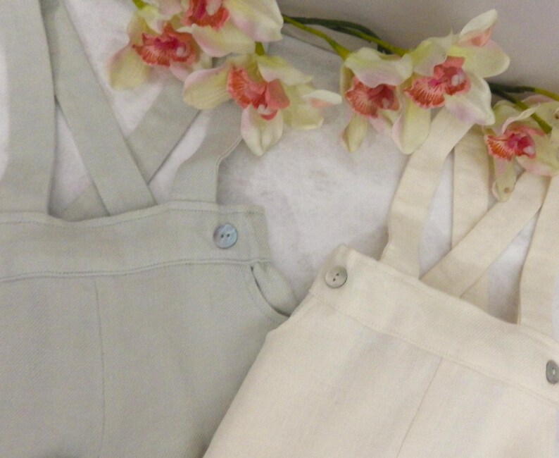 May include: Two pairs of linen overalls, one in a light green color and the other in a cream color. Both pairs have adjustable straps and button closures.