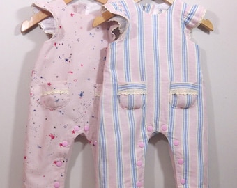 May include: Two baby rompers hanging on wooden hangers. One is pink with a celestial print, the other is striped in pink and blue. Both have ruffled sleeves, lace-trimmed pockets, and pink button closures.