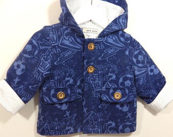 May include: A blue hooded jacket with a sports-themed pattern, featuring soccer balls, sneakers, and the words "COOL" and "YEAH". The jacket has wooden buttons and white cuffs. The label reads "cara secco".