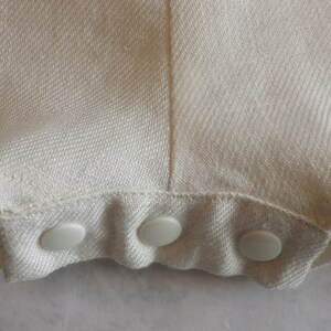 May include: Close-up of a white fabric garment with a row of four white plastic buttons along the bottom edge.