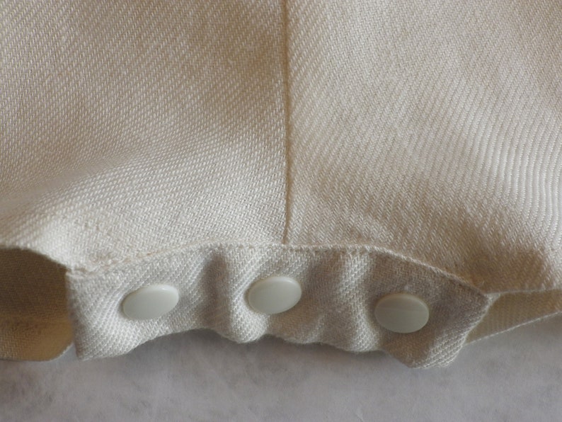 May include: Close-up of a white fabric garment with a row of four white plastic buttons along the bottom edge.