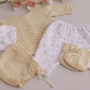 May include: A set of knitted baby clothes in a cream colour, including a sweater, trousers, booties, and a hat. The trousers have a white background with a small brown print.