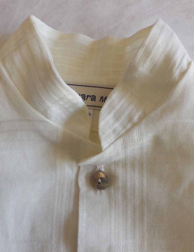 May include: White linen shirt with a mandarin collar and a single button closure. The shirt has a textured weave and a label that reads "Mara ne".