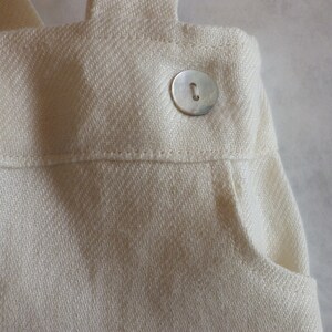 May include: A close-up of a white cotton fabric with a single silver button. The fabric has a subtle herringbone pattern and a pocket on the right side.