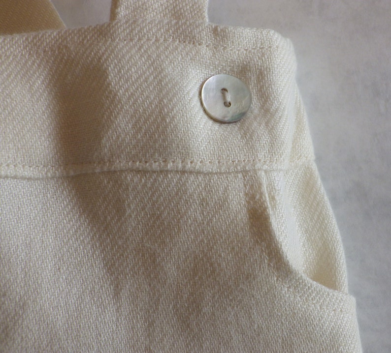 May include: A close-up of a white cotton fabric with a single silver button. The fabric has a subtle herringbone pattern and a pocket on the right side.