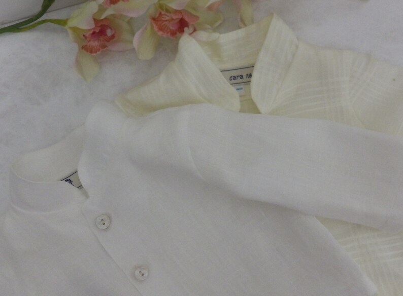 May include: Two white linen shirts with button closures. The shirt on top has a collar and a label that reads "cara ma".