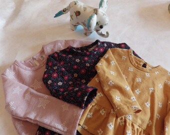 May include: Three long-sleeved shirts in pink, black with white floral print, and yellow with white animal print. The shirts are folded and stacked on top of each other.