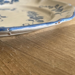 May include: A cream-colored ceramic plate with a scalloped edge and a blue painted floral pattern. The rim features a blue painted border with visible signs of wear, including small chips and areas where the glaze has worn away along the edge.