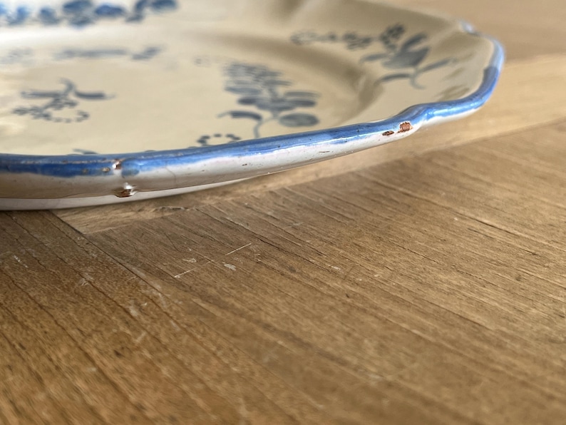May include: A cream-colored ceramic plate with a scalloped edge and a blue painted floral pattern. The rim features a blue painted border with visible signs of wear, including small chips and areas where the glaze has worn away along the edge.