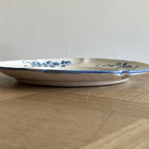 May include: A round ceramic plate with a cream-colored base and a hand-painted blue floral pattern. The plate features a scalloped edge with a thin blue rim and visible fine crackle glazing across the surface, viewed from a low side angle.