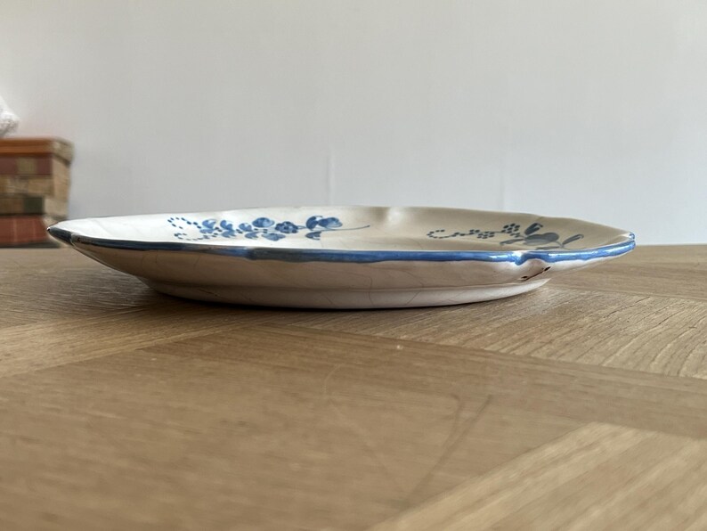 May include: A round ceramic plate with a cream-colored base and a hand-painted blue floral pattern. The plate features a scalloped edge with a thin blue rim and visible fine crackle glazing across the surface, viewed from a low side angle.
