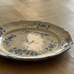 May include: A round ceramic plate with a scalloped edge, featuring a cream-colored base and a hand-painted blue floral pattern consisting of delicate vines, leaves, and small blossoms arranged in a circular design around the center.