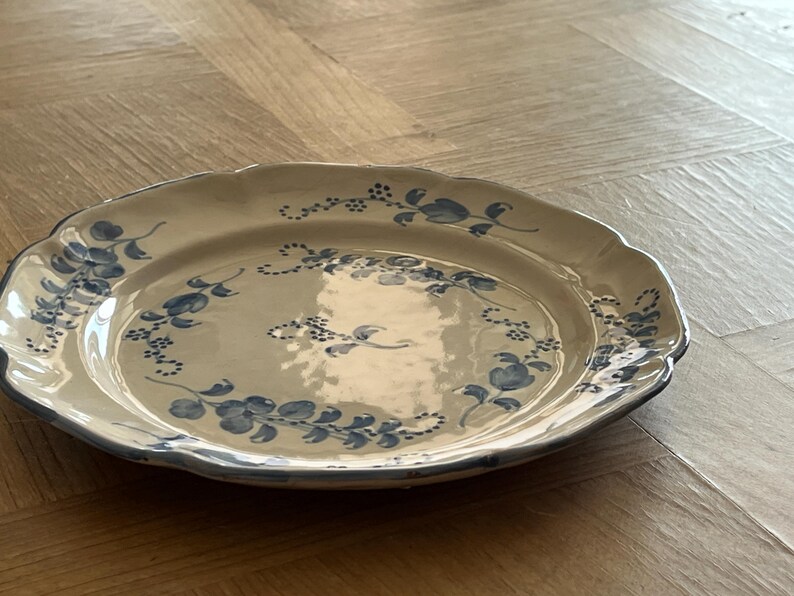 May include: A round ceramic plate with a scalloped edge, featuring a cream-colored base and a hand-painted blue floral pattern consisting of delicate vines, leaves, and small blossoms arranged in a circular design around the center.